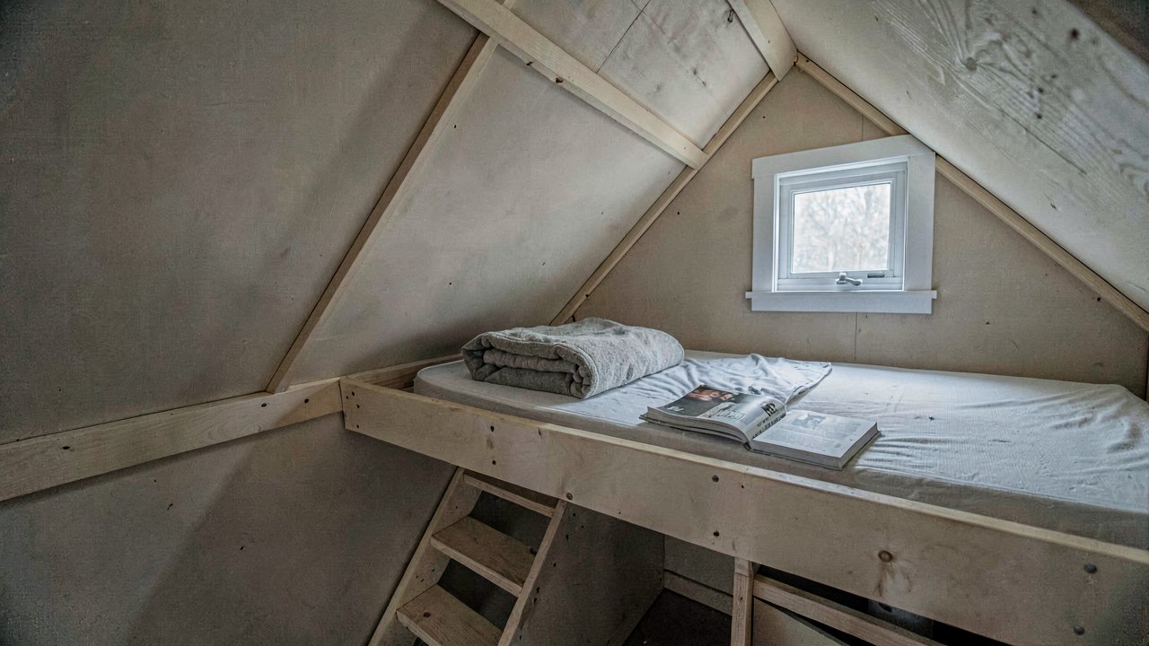 Tiny house sleeping loft with a mattress under a low sloped ceiling and a small gable window